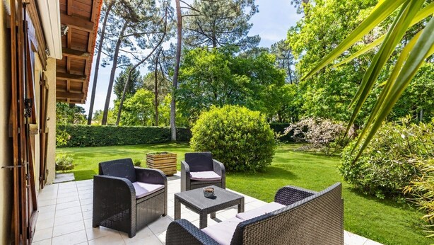 Een terras met rieten meubels, een houten tafel en uitzicht op een verzorgde tuin met bomen en struiken.