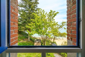 Une vue à travers une fenêtre, avec un mur de briques et un arbre au premier plan, qui s’ouvre sur une plage.