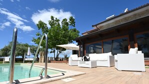 Zona junto a la piscina con pileta, mobiliario de mimbre blanco y un ventanal con vista a la piscina.