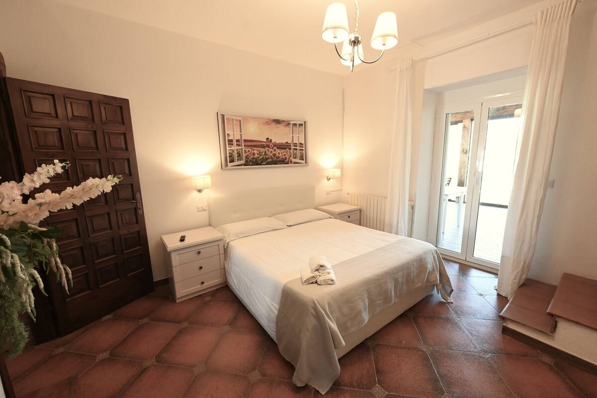 Une chambre à coucher avec un lit, des tables de chevet, un tableau accroché au mur, une porte et une vue à travers une porte vitrée.