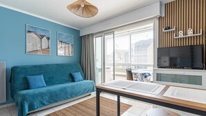 Ein modernes Wohnzimmer mit einem türkisblauen Sofa, einem Esstisch mit Untersetzern, einem Fernseher und einem großen Fenster mit Jalousien.