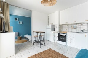 A compact kitchen with white cabinets, a stove, and a small dining area with stools. - Studio Côte de Nacre - À 450 m de la Plage (Courseulles-sur-Mer)