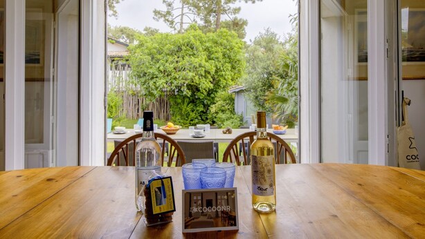 A wooden dining table with a bottle of wine, a framed picture, and a blue glass set. - Villa Veronica - À 50 m de la plage (Lège-Cap-Ferret)
