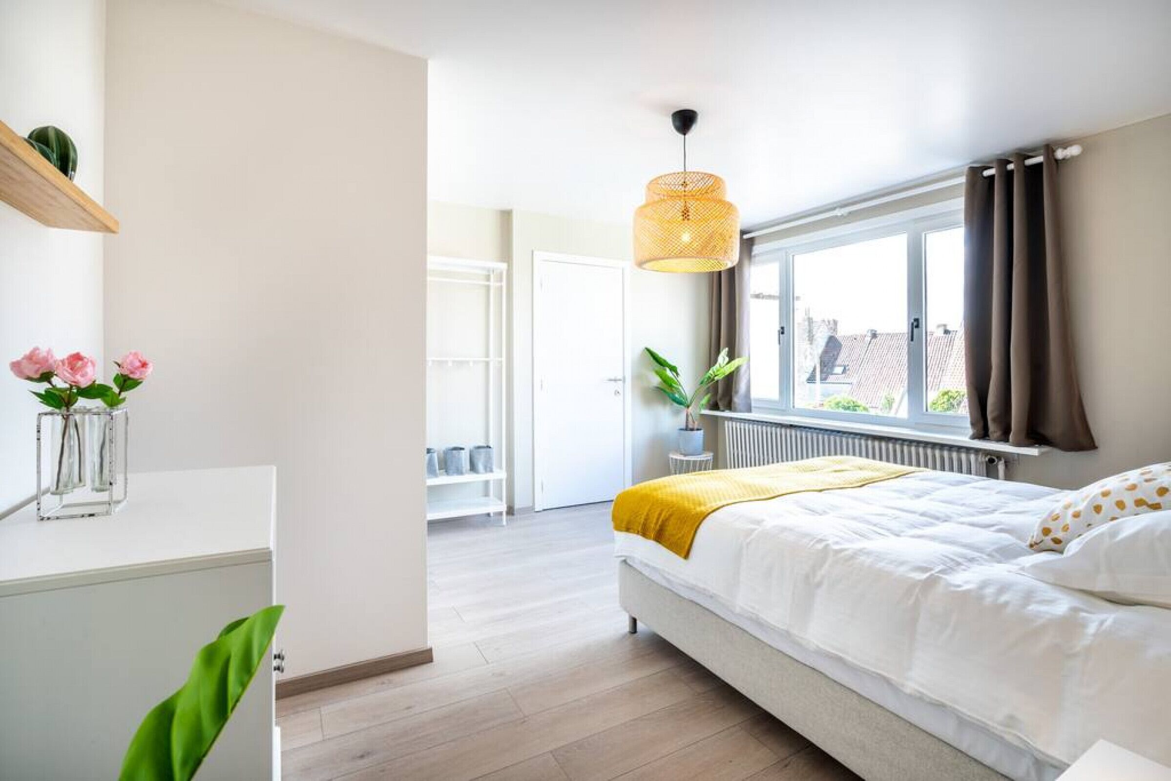 Ein Schlafzimmer mit Bett, einem Fenster mit Vorhängen, einem Nachttisch mit Pflanze und einer Hängelampe.