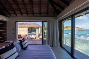 Une chambre avec un grand lit, une tête de lit en bois, une vue sur l’océan et un mur en pierre.