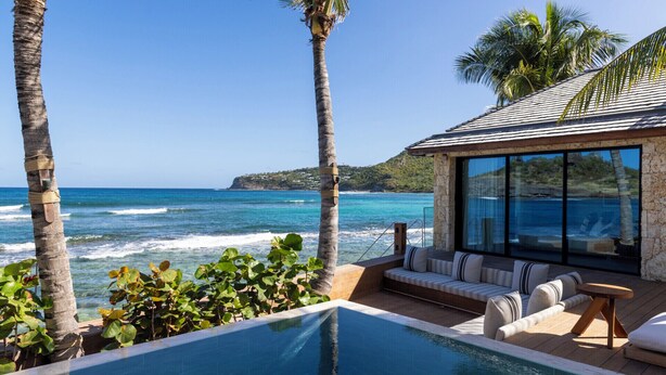 A poolside area with a view of the ocean, a stone building with large windows, and a seating area with cushions.