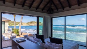 A bedroom with a large window overlooking the ocean, a wooden ceiling, and a bed with white linens.