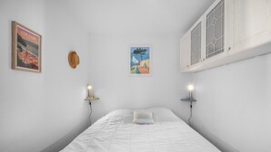 Quarto com cama, duas luminárias fixadas na parede, um cartaz emoldurado e janela com vista.