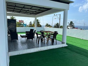 A covered outdoor patio with a table and chairs, a grill, and a view of the ocean.