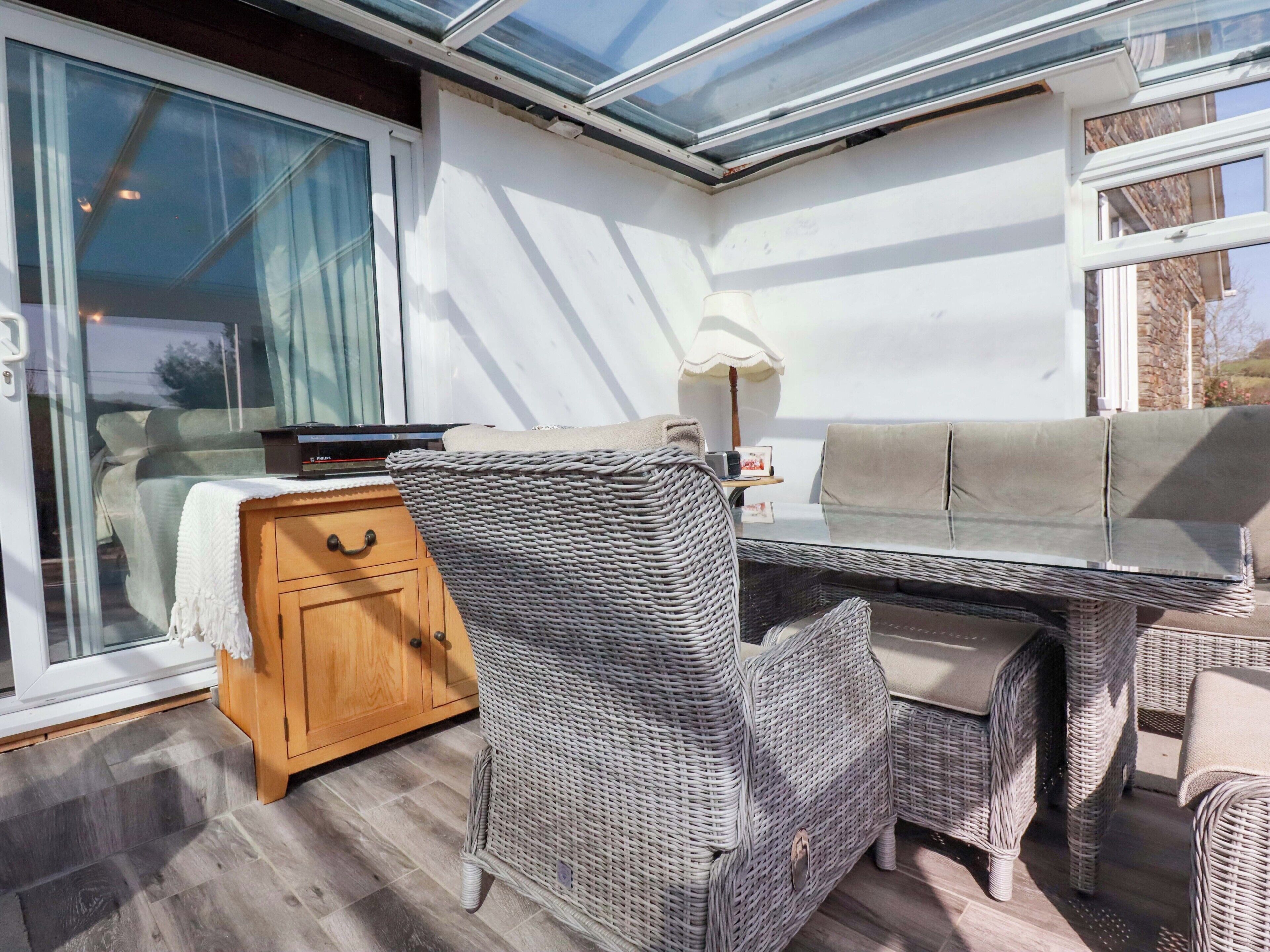Una habitación iluminada por el sol con techo de vidrio, muebles de mimbre, un aparador de madera y una mesa con lámpara.