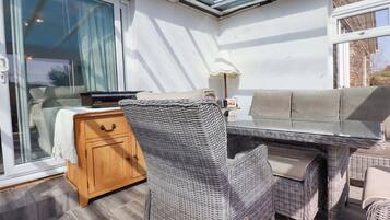 Una habitación iluminada por el sol con techo de vidrio, muebles de mimbre, un aparador de madera y una mesa con lámpara.