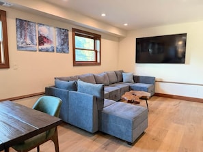A living room with a sectional sofa, a coffee table, and a dining table. There are three framed pictures on the wall and a large TV mounted on the wall. - Historic Stone Villa | Donner | Sauna | EV Charger (Soda Springs)