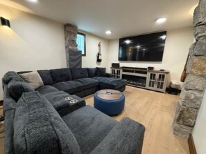 A modern living room with a sectional sofa, a round ottoman, a flat-screen TV, and a stone accent wall. - Historic Stone Villa | Donner | Sauna | EV Charger (Soda Springs)