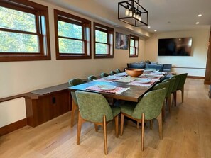 A dining area with a long wooden table, green upholstered chairs, and a large window with a view of trees.