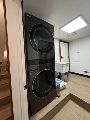 A modern laundry room with a black wall-mounted washer and dryer, a wooden bench, a white sink, and a tiled floor.