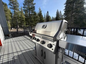 Una barbacoa d'acer inoxidable per a l'exterior amb quatre cremalls, situada sobre un terrat de fusta i barana metàl·lica.
