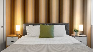Un lit bien fait, avec une tête de lit grise, deux tables de chevet avec des lampes et un mur en lambris de bois derrière.