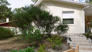 A house with a beige exterior, a small garden with various plants, and a set of beige steps leading to the front door. - Single-storey house under the pines in a peaceful setting between lake and ocean (CARCANS)