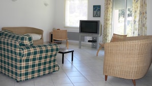 A living room with a sofa, a chair, a television on a stand, and a window with curtains. - Single-storey house under the pines in a peaceful setting between lake and ocean (CARCANS)