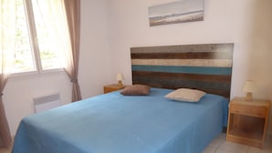 A bedroom with a bed, a nightstand, a lamp, a window with curtains, a radiator, and a painting on the wall. - Single-storey house under the pines in a peaceful setting between lake and ocean (CARCANS)