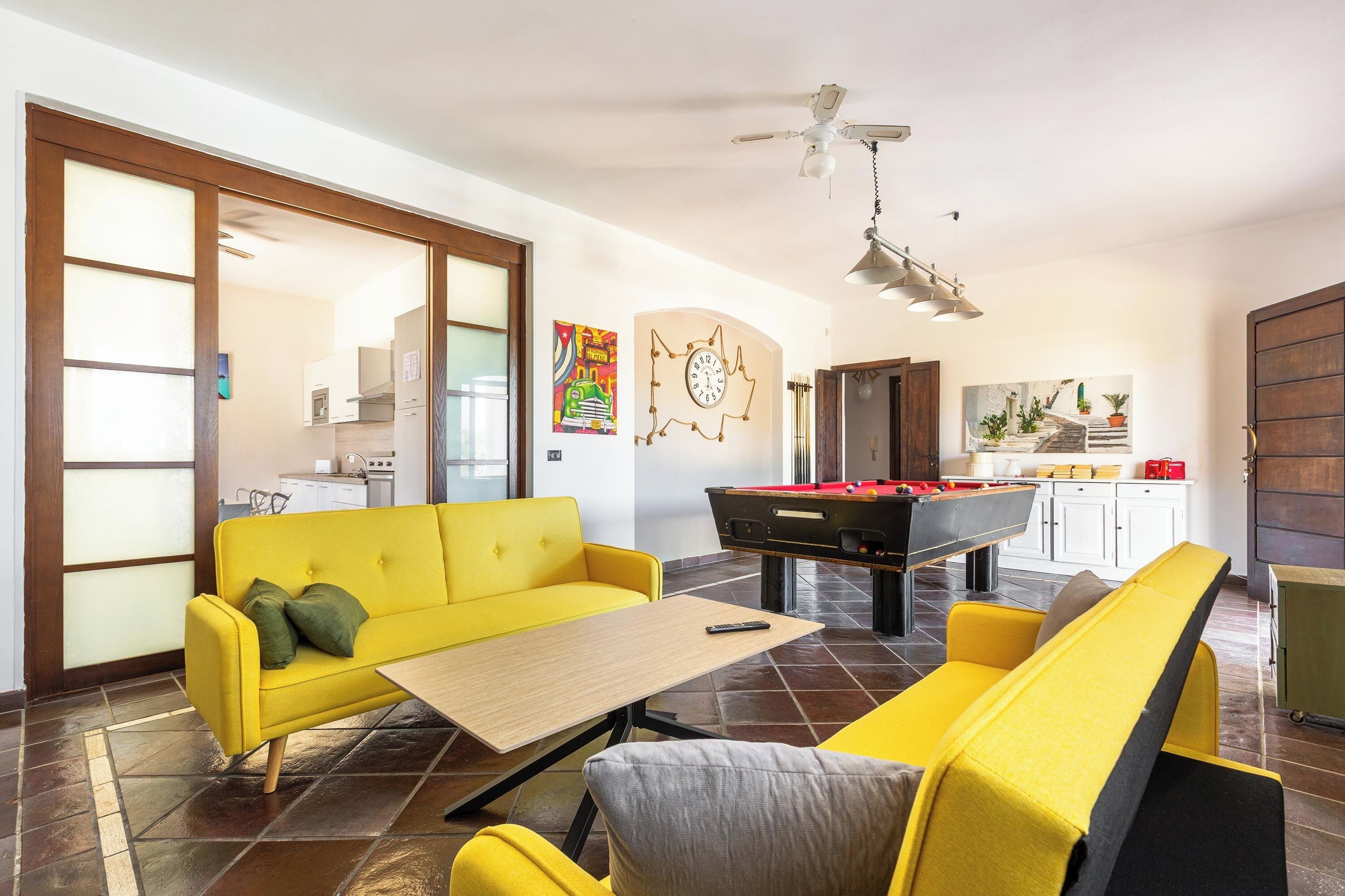 Un salon avec un canapé jaune, une table de billard et une cuisine à l’arrière-plan.