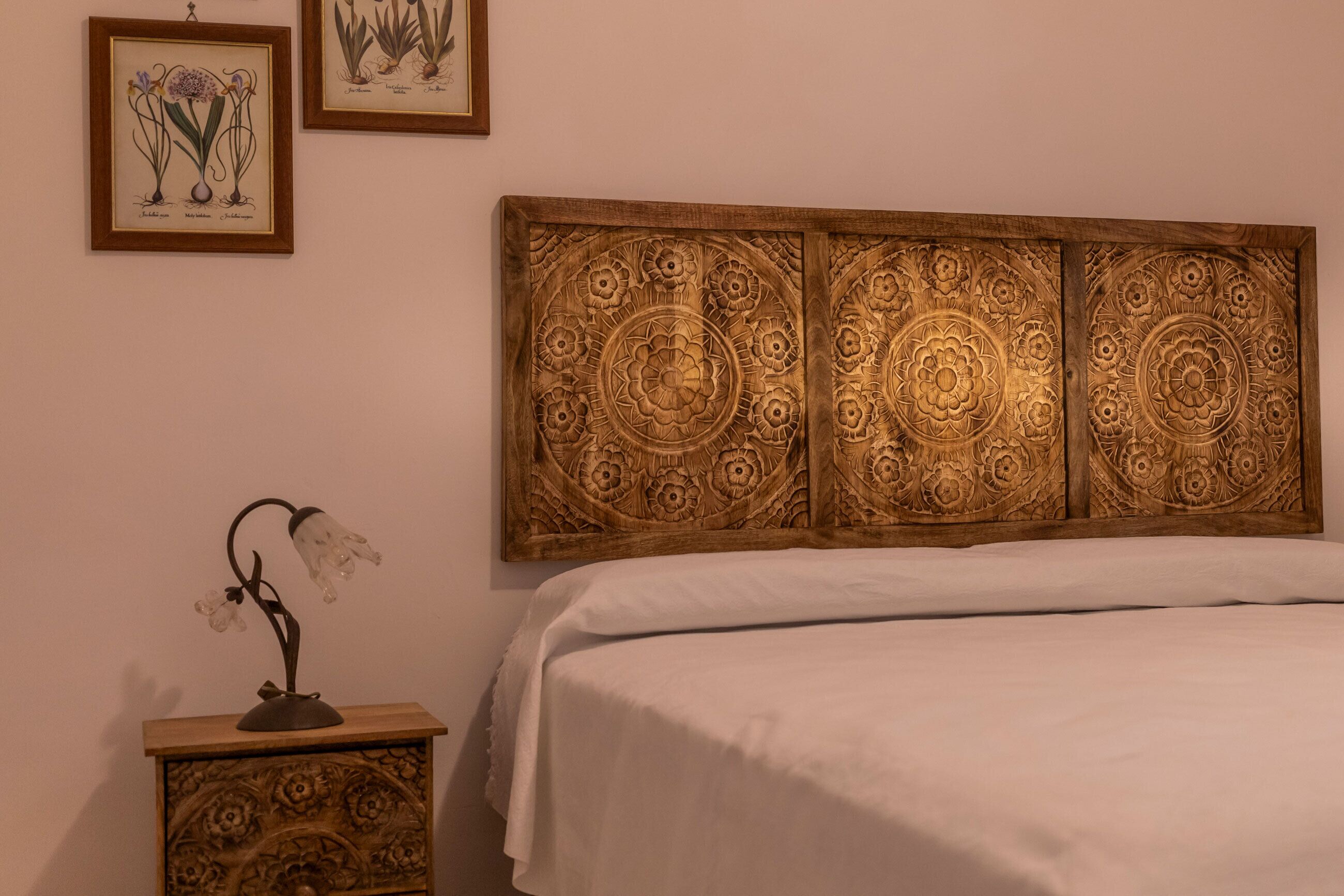 Un dormitorio con una cama, una mesita de noche con lámpara y una pared decorada con ilustraciones botánicas enmarcadas.