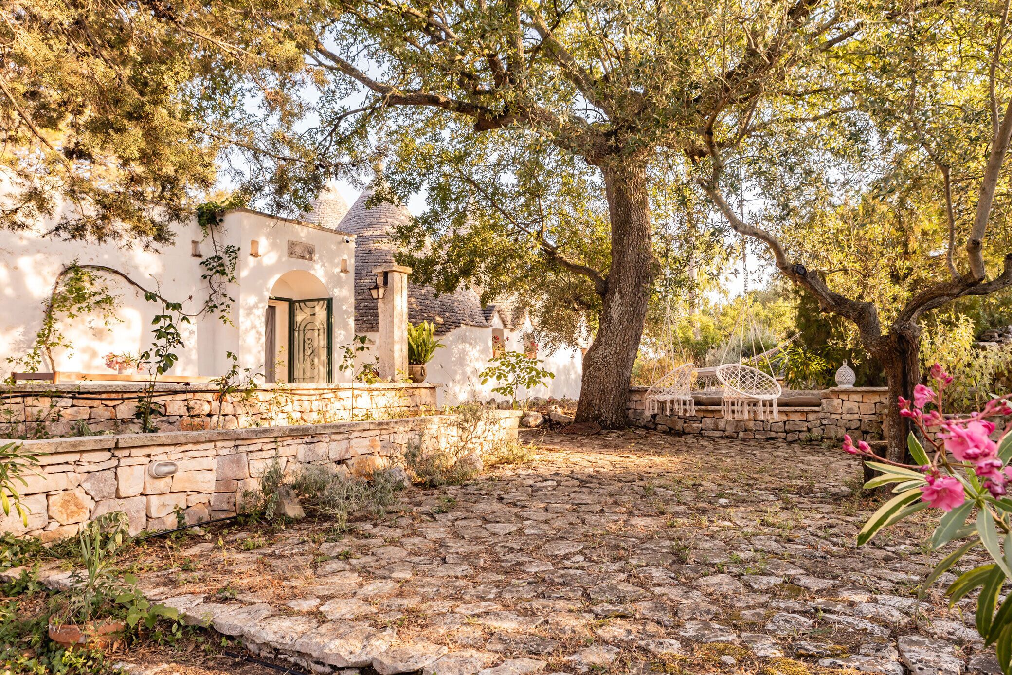 A stone-paved courtyard with a large tree, stone walls, and a white building with an arched doorway.