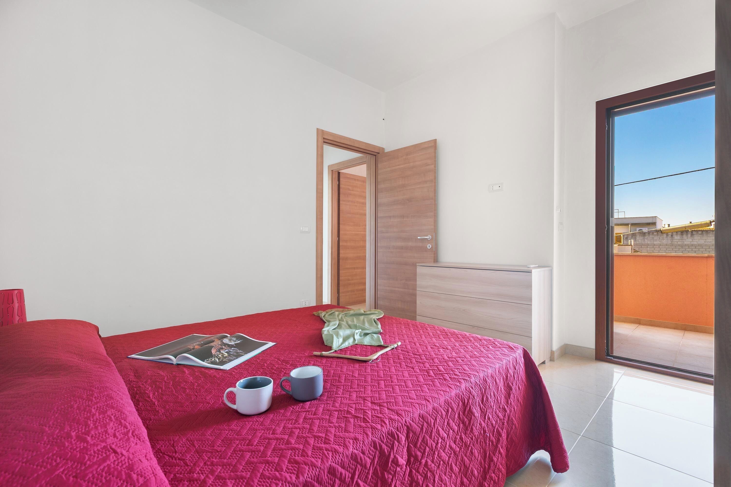 A bedroom with a red bedspread, a white bedside table, and a view through a sliding glass door to an outdoor balcony.