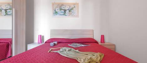 A neatly made bed with a red quilt, a beige dress, and a magazine on it. There are two bedside tables with pink lamps and a framed painting on the wall.