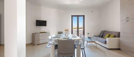 A modern living room with a dining area, a sofa, a TV mounted on the wall, and a large window.