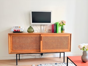 A wooden cabinet with a television on top, books, a vase with flowers, and a small table with a vase. - Heart Marais Studio by Melvane (Paris)