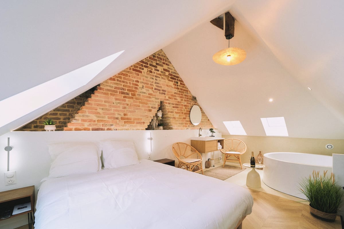 A bedroom with a sloped ceiling, a brick wall, a large bed, a sitting area with chairs, a vanity, and a bathtub.