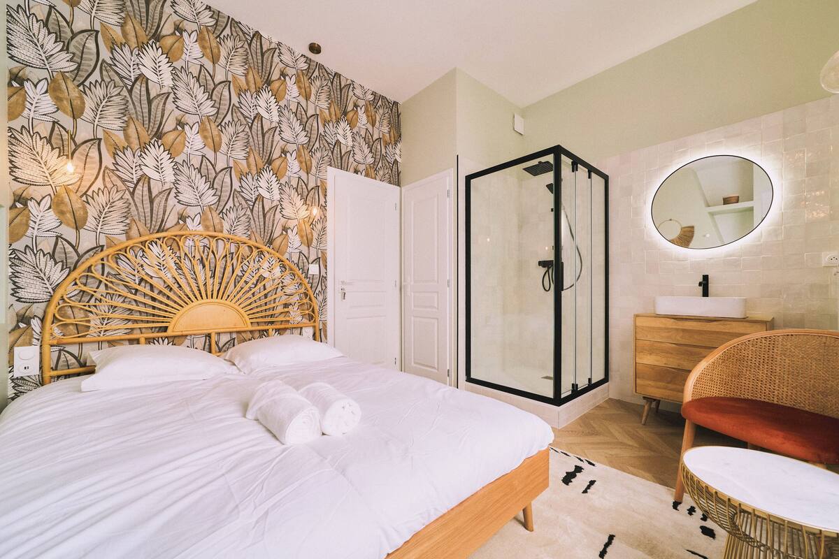 A bedroom with a bed, a shower, a vanity with a mirror, and a rattan chair.