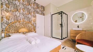 A bedroom with a bed, a shower, a vanity with a mirror, and a rattan chair.