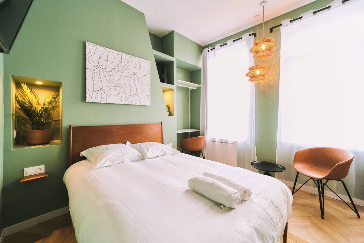 A neatly made bed with white linens, a wooden headboard, and a rolled towel on top. There is a single chair and a small table beside the bed.