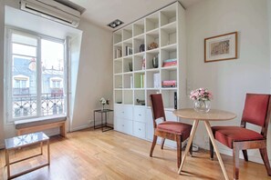 Una habitación con piso de madera, una estantería blanca, una mesa redonda con sillas, un banco y un ventanal con vista a edificios.