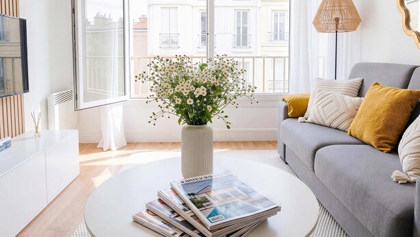Een moderne woonkamer met een ronde salontafel, een grijze bank, een witte vaas met bloemen en een stapel tijdschriften.