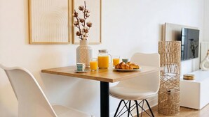 Een moderne eetruimte met een houten tafel, witte stoelen, een vaas met gedroogde bloemen, een glas sinaasappelsap, een pot sinaasappelsap en een bord croissants.