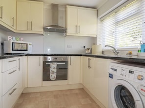 A kitchen with a microwave, oven, and washing machine, cabinets, and a window with blinds.