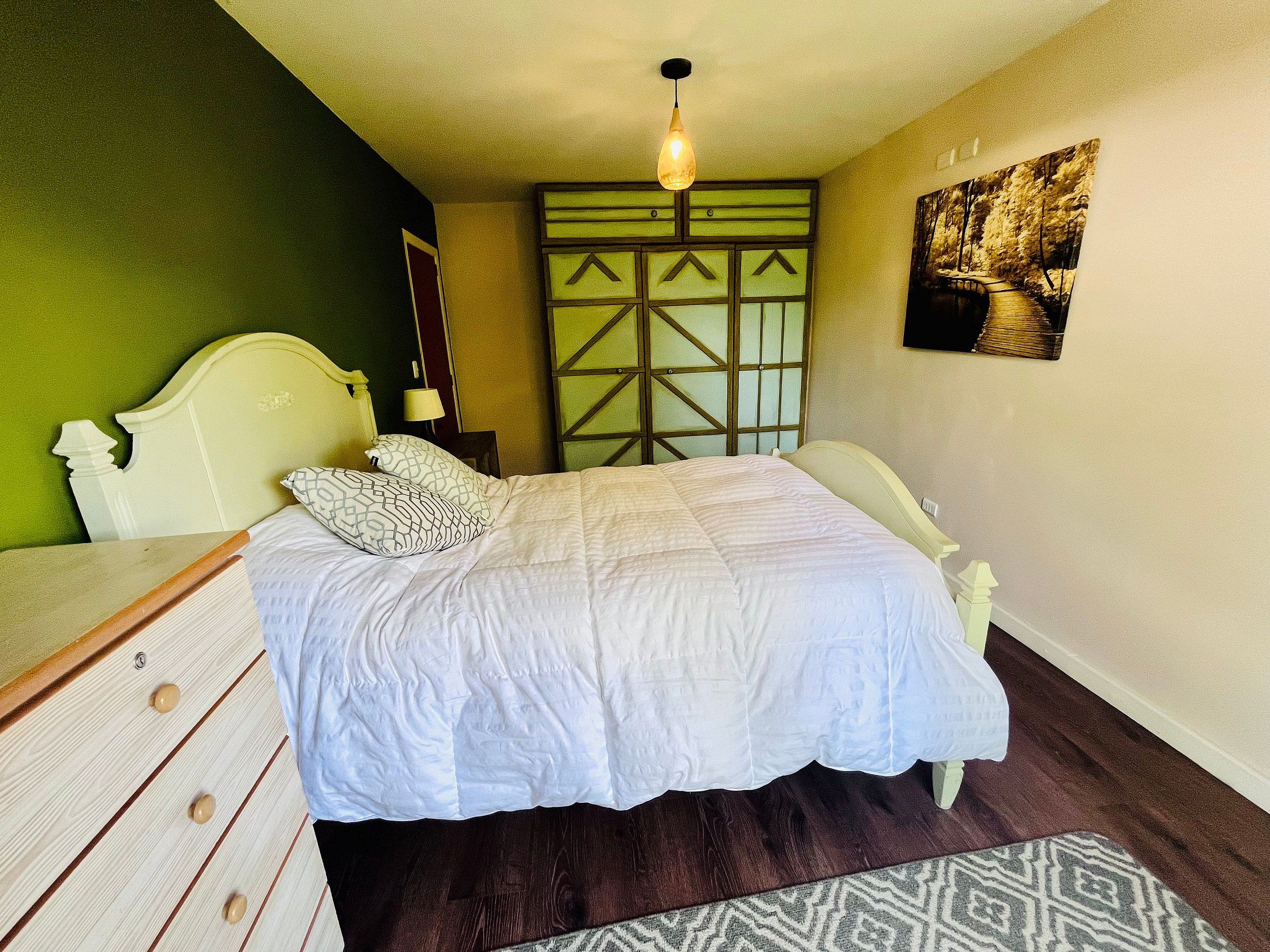 Un dormitorio con cama, cómoda, mesita de noche, lámpara y un cuadro en la pared.