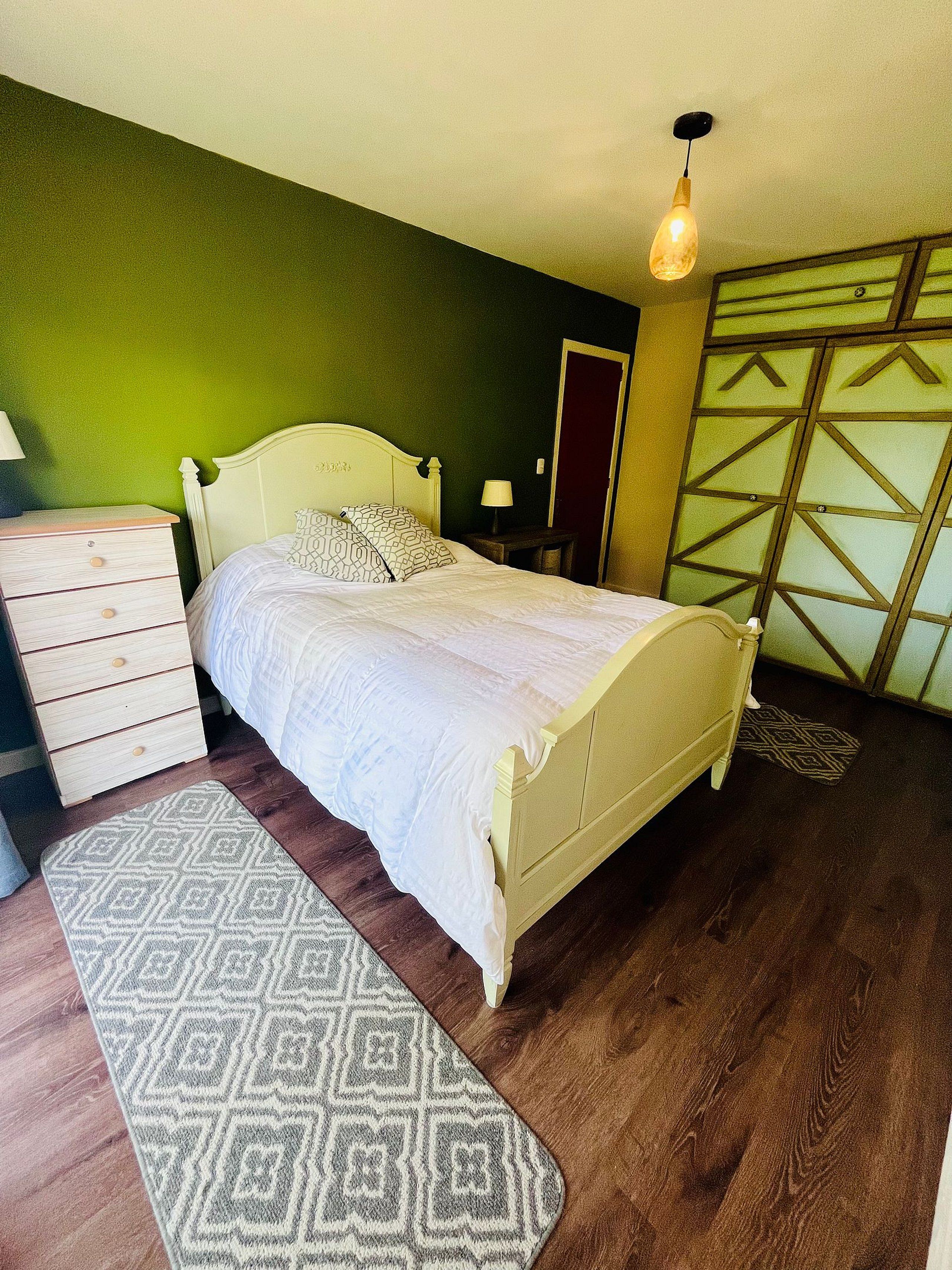 A bedroom with a bed, dresser, nightstand, lamp, and patterned rug.