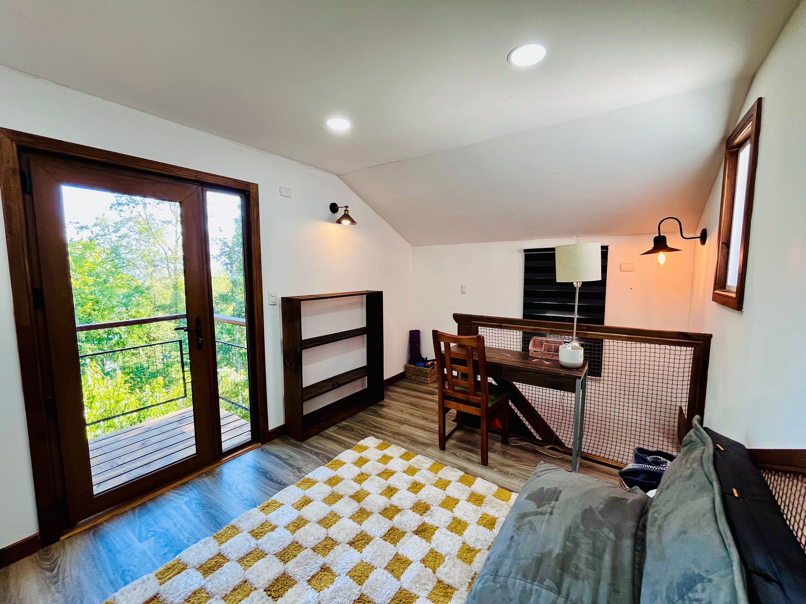 Quarto com mesa de jantar, cadeiras, um banco, sofá, tapete estampado e vista para uma varanda externa através de uma porta.