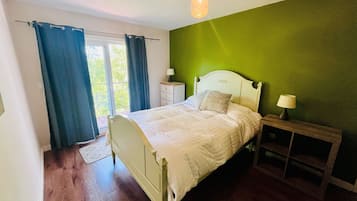 Un dormitorio con cama, mesita de noche, lámpara, ventana con cortinas y vista a zonas verdes.
