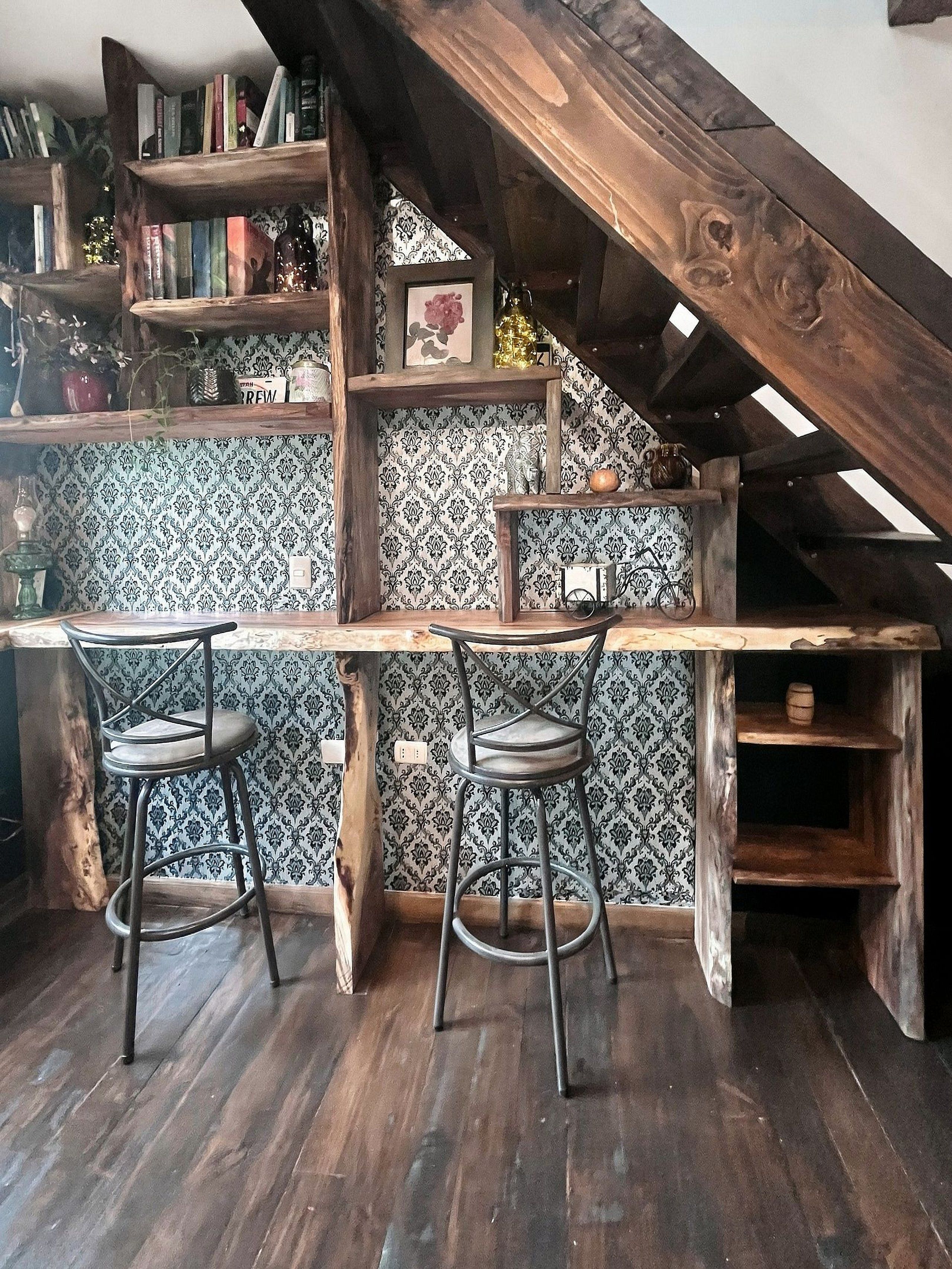 Um espaço de bar rústico com móveis de madeira, dois banquetas e uma estante cheia de livros e objetos decorativos.
