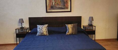 A large bed with a dark blue bedspread, two pillows, and a framed artwork above it. There are two bedside tables with lamps on either side of the bed.