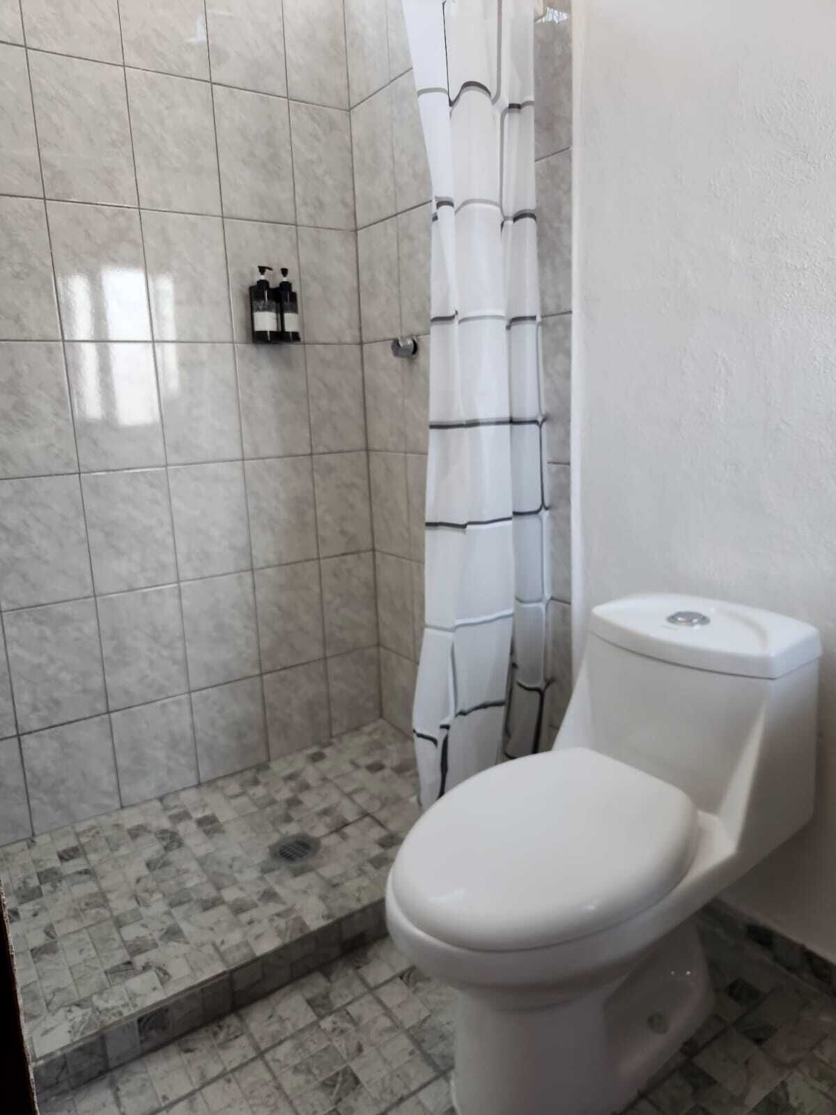 Un baño con inodoro, ducha con cortina blanca y suelo de baldosas.