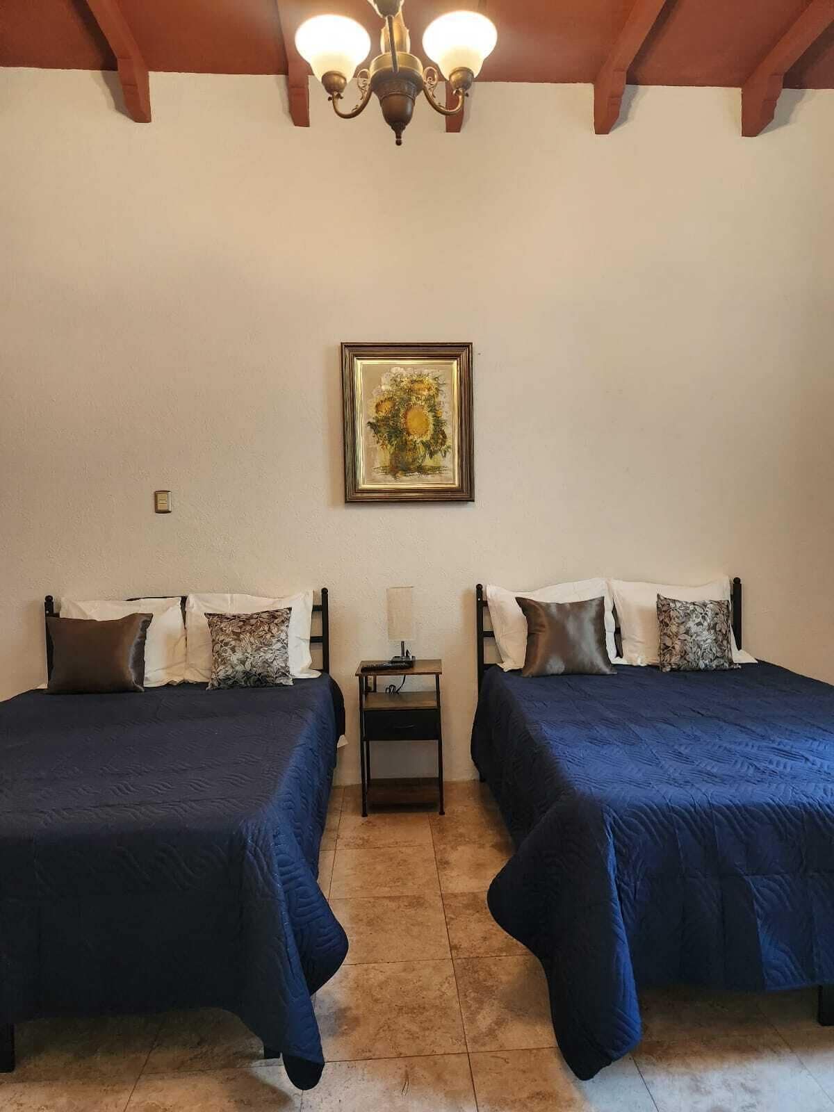 Una habitación con dos camas, una mesita de noche, un cuadro enmarcado en la pared y una lámpara en el techo.