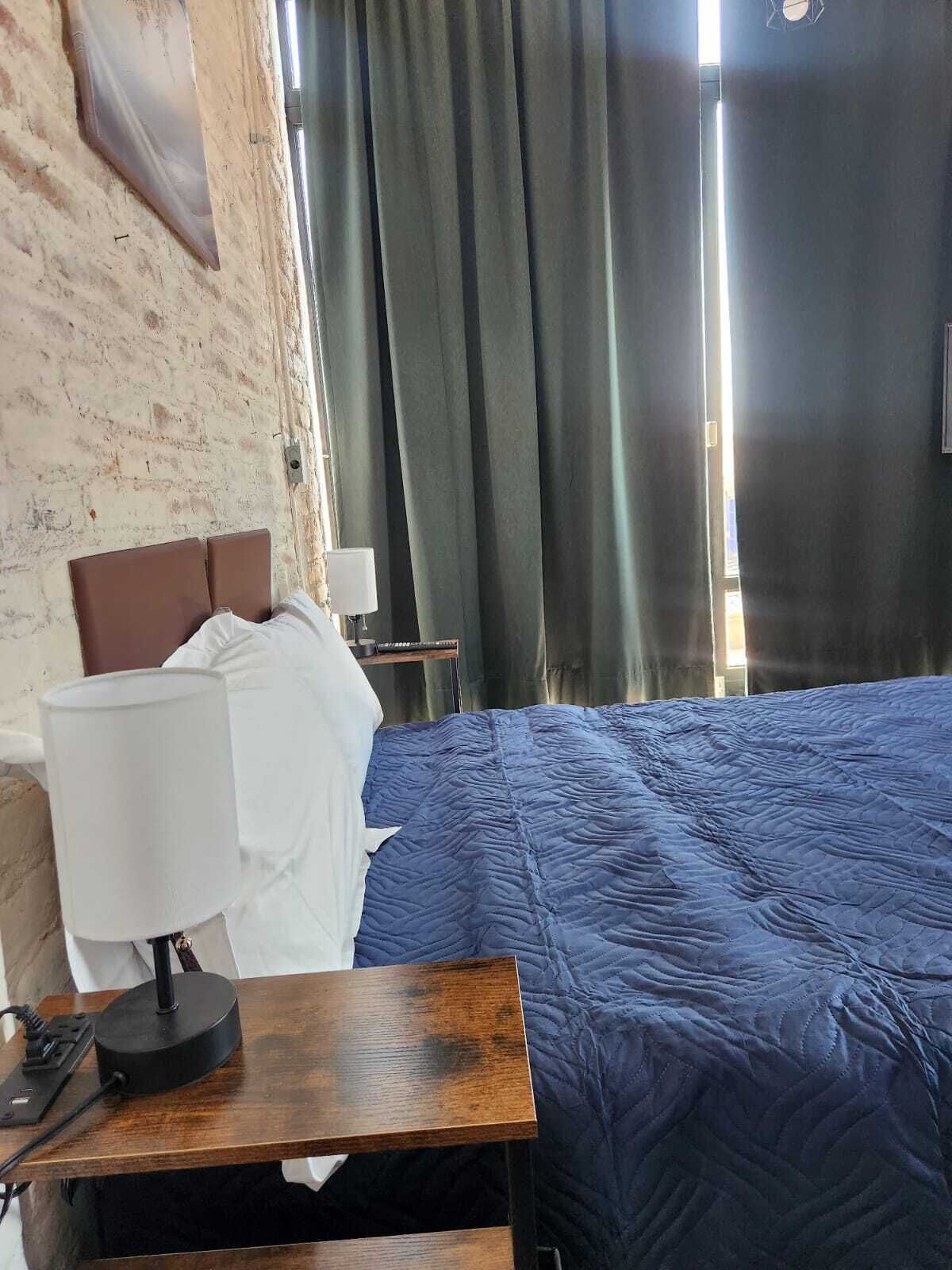 Habitación de hotel con cama, mesita de noche de madera con lámpara, pared de ladrillo y ventana con cortinas.