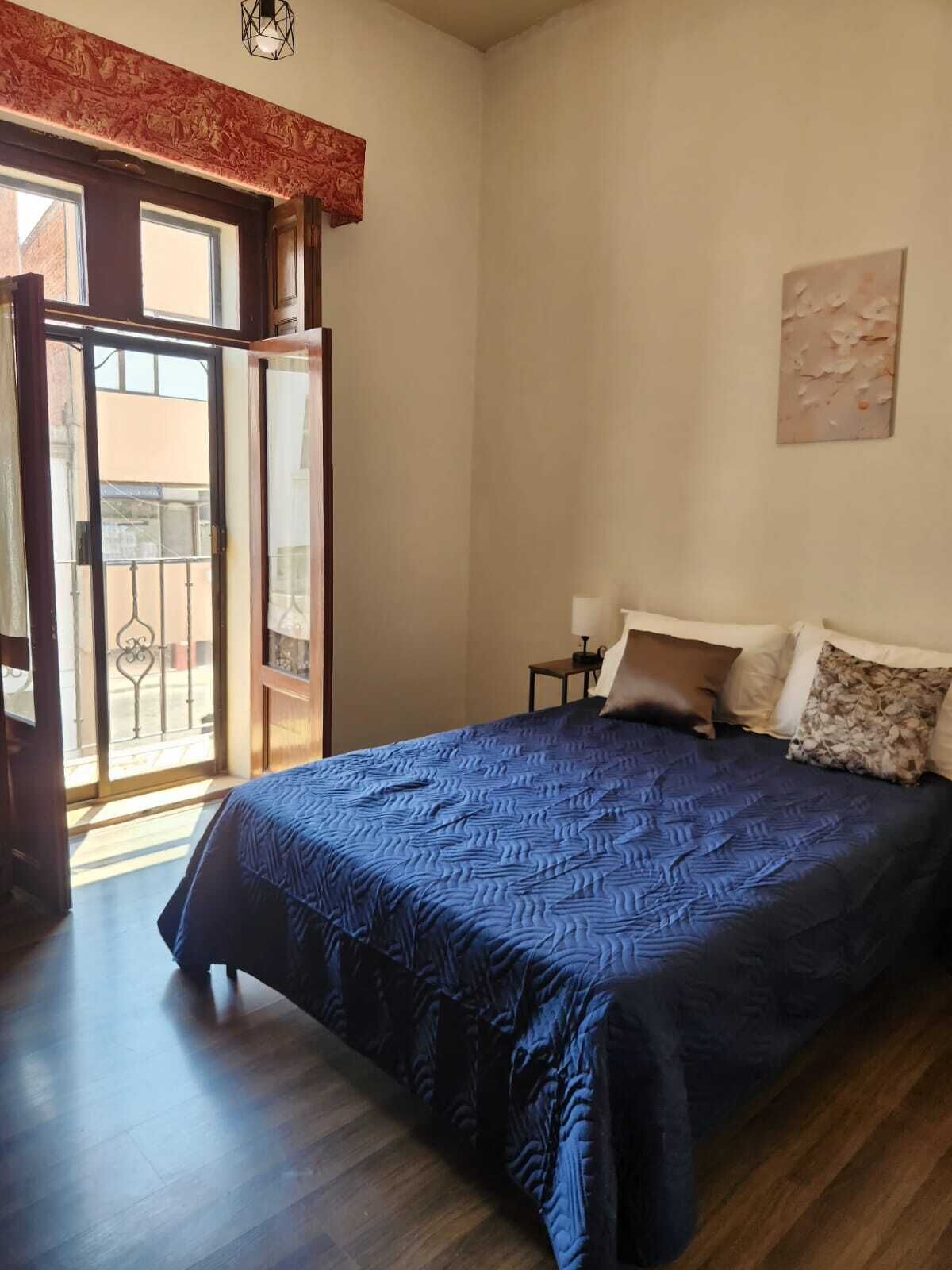 Un dormitorio con una cama grande, una mesita de noche, una lámpara, un cuadro en la pared y un ventanal corredero que da a un balcón.