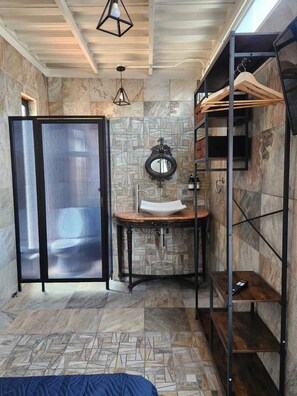 A bathroom with a stone wall, a sink, a mirror, and a shower area. - Hotel Casa Victoria (Queretaro)